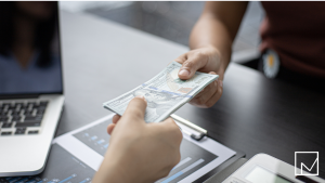 Businessperson handing money to another across a table, symbolizing how staffing agencies earn revenue when first starting out.