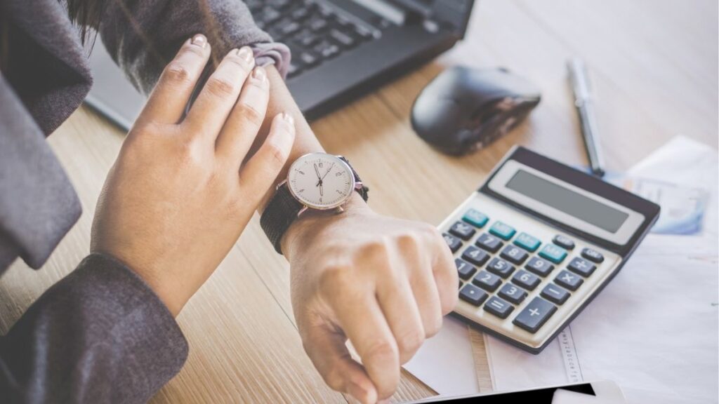 Professional checking watch beside calculator and laptop, representing time management and financial planning for staffing agency operations.