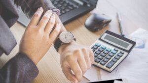 Professional checking watch beside calculator and laptop, representing time management and financial planning for staffing agency operations.