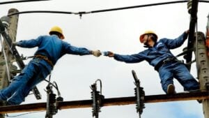 Two utility workers exchanging equipment on a power line, representing workplace risk, safety, and workers’ compensation considerations for staffing firms.