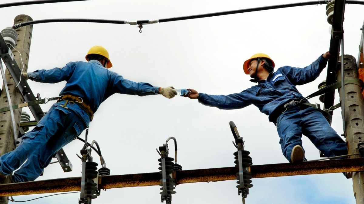 Two utility workers exchanging equipment on a power line, representing workplace risk, safety, and workers’ compensation considerations for staffing firms.