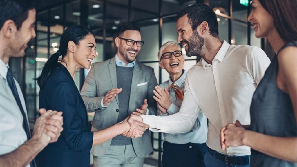 Business professionals shaking hands in an office while team celebrates a successful partnership, representing payroll funding support for staffing firms.