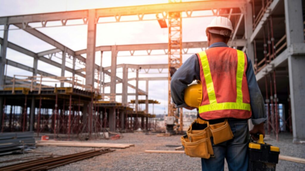 Construction worker in safety vest and hard hat walking through active jobsite with steel framework and crane in background.