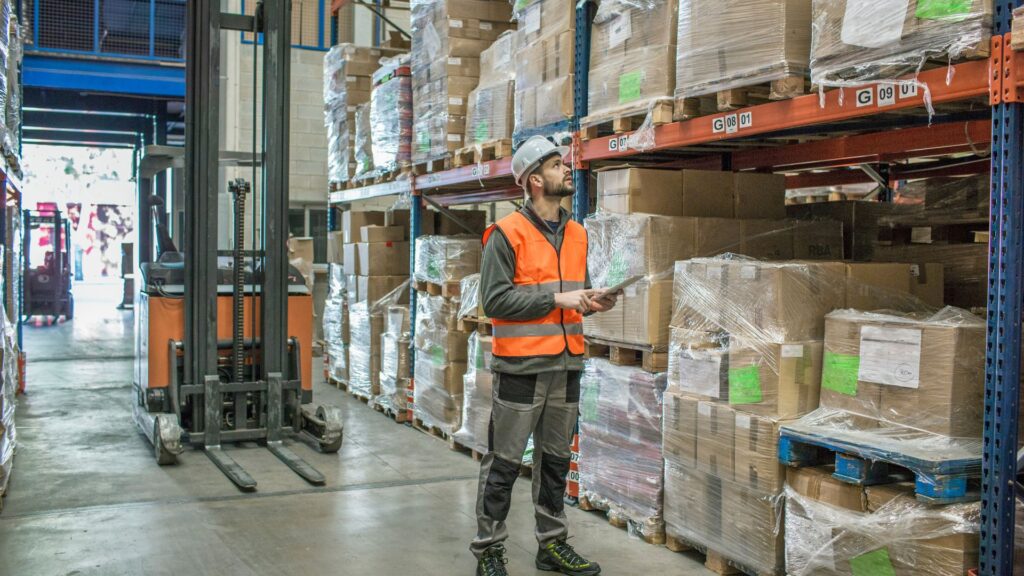 Light industrial warehouse worker conducting inventory inspection near forklift and palletized shipments in distribution facility.