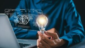 Person holding a glowing light bulb in front of a laptop with a digital AI brain interface, representing AI adoption in staffing, innovation, technology implementation, and building trust in artificial intelligence solutions.