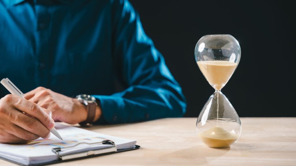 Person in a blue shirt writes on papers at a desk, with an hourglass timer in the foreground on the right.