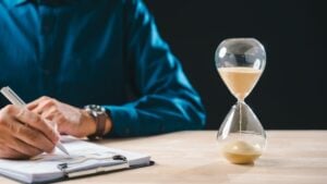 Person in a blue shirt writes on papers at a desk, with an hourglass timer in the foreground on the right.