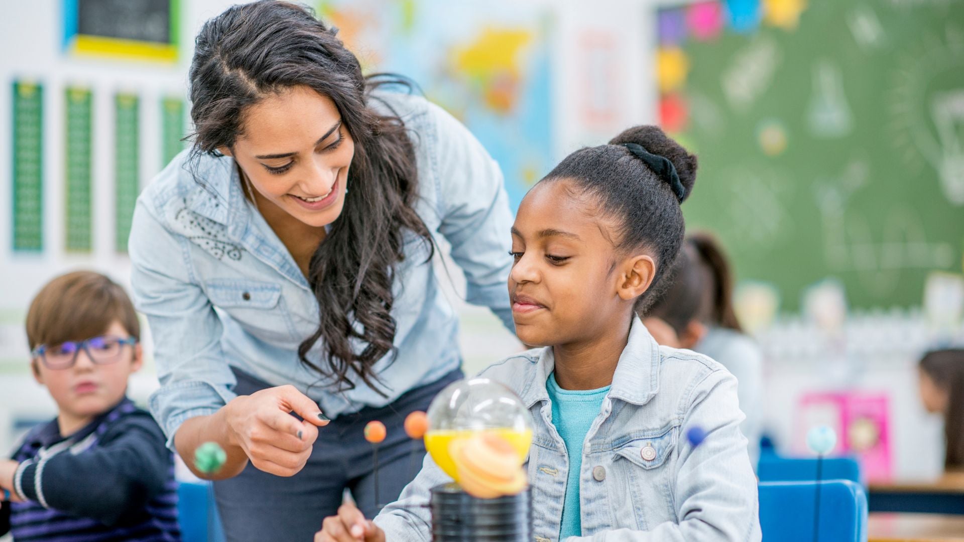 Teacher helping an elementary student with a solar system model in a classroom.